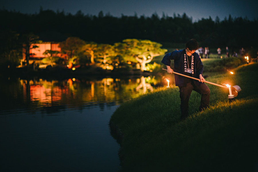 Worker lighting torches along the edge of a Japanese garden pond at night with illuminated trees