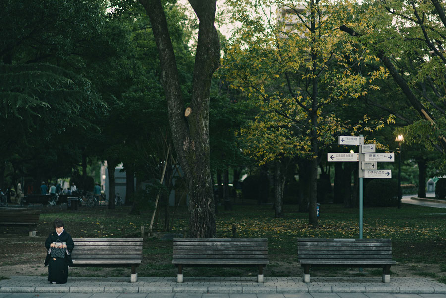 Person sitting alone on a park bench under large trees with autumn leaves beginning to turn