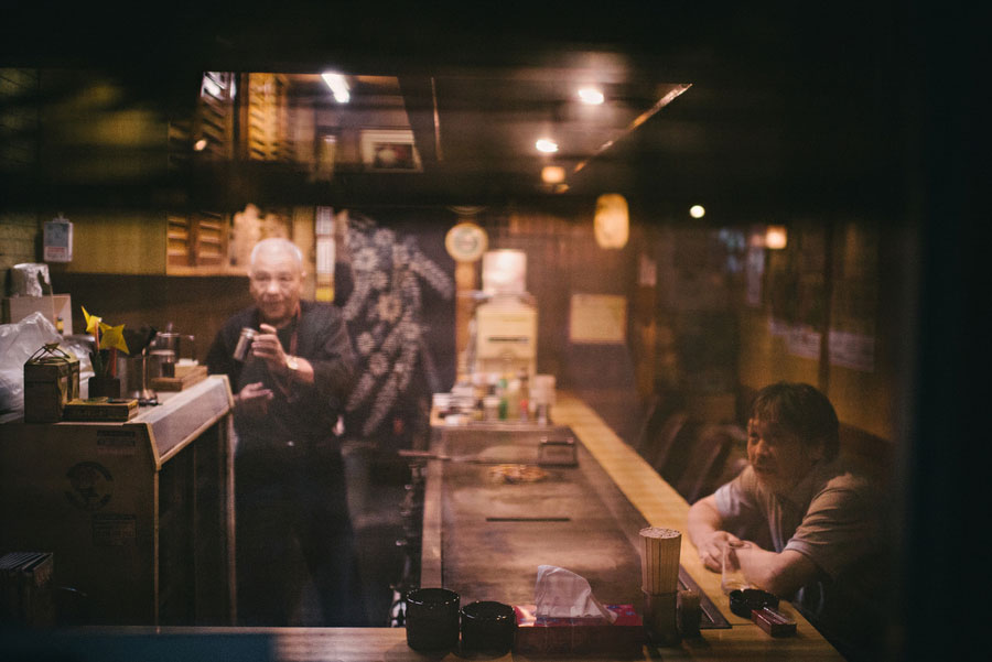Two men sitting at a dimly lit izakaya counter seen through a window reflection at night
