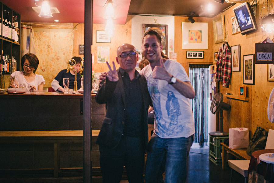Two men posing with peace signs in the doorway of a small Japanese bar at night