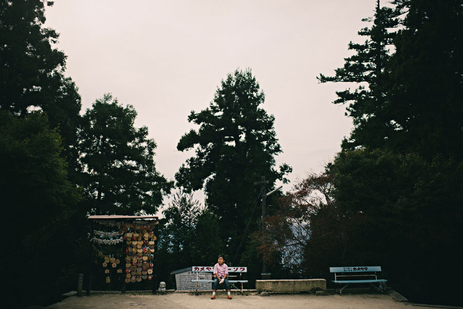 Child sitting alone on a stone platform between tall cedar trees at a shrine entrance