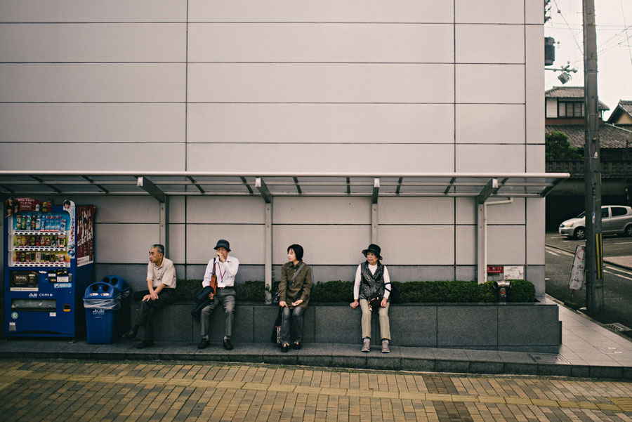 Four people sitting spaced apart on a bench outside a modern building beside a vending machine