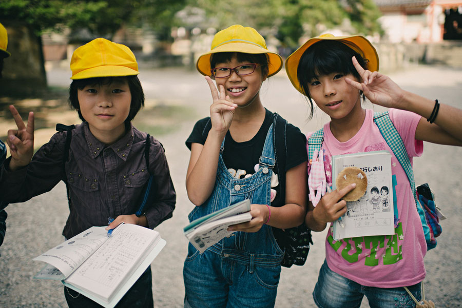 Three schoolgirls in yellow hats holding notebooks and snacks posing with peace signs