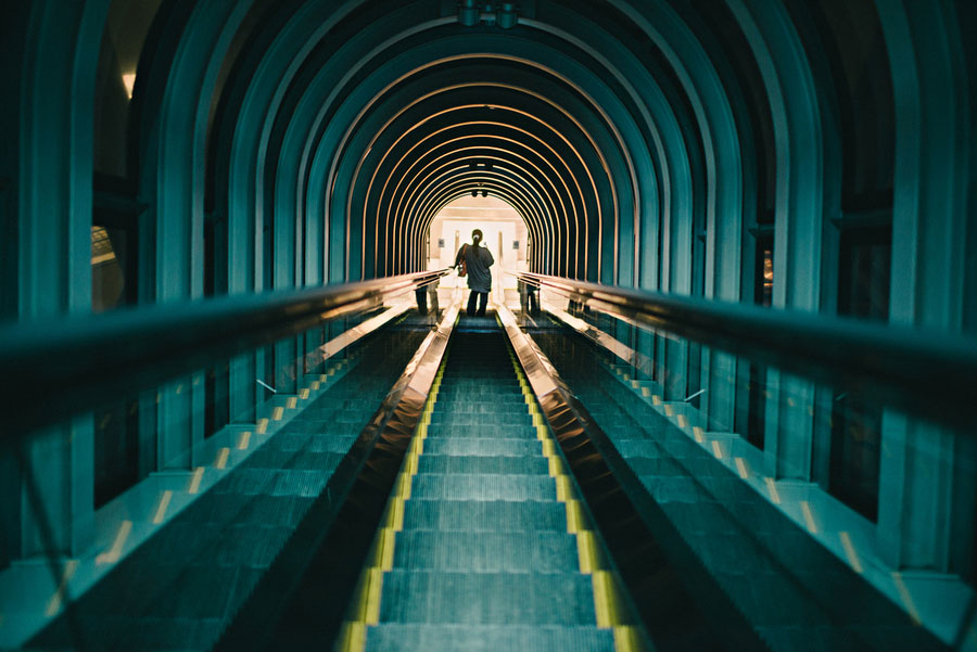 Person silhouetted at the top of a long escalator tunnel with concentric teal arches