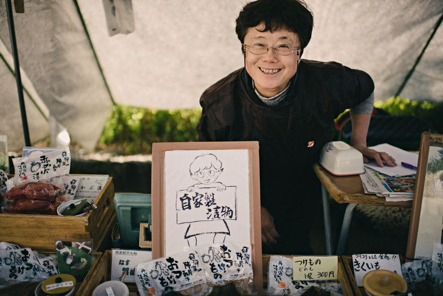 Smiling woman behind a market stall holding a hand-drawn self-portrait sign selling homemade pickles