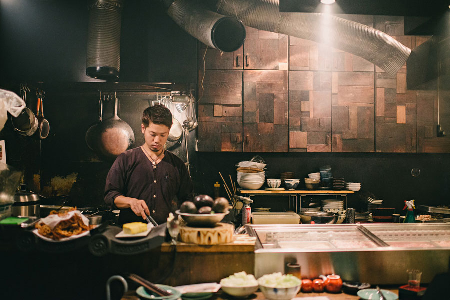 Chef preparing food at a rustic open kitchen counter in a warmly lit Japanese restaurant